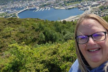 Tammy Spantiko smiles on a hill in front of a forest with a harbor and seaside city in the background.