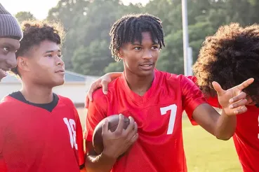 Four teen boys stand together smiling on a school football field. One is holding a football and pointing across the field.