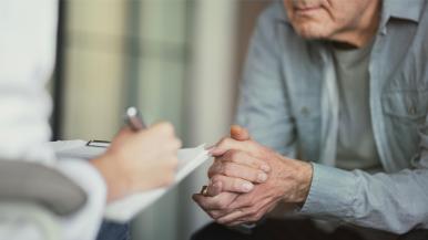 Older man clasps his hands as another person's hands are shown writing on a clipboard