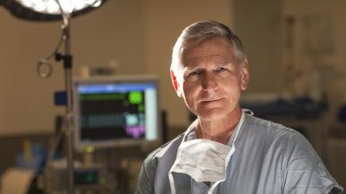 Dr. Fessler in surgical scrubs with a face mask around the neck, standing in a clinical setting with medical equipment and a monitor in the background.