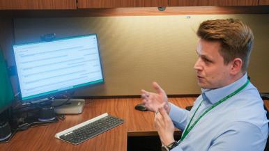 A man seated at a wooden desk gesturing toward a computer monitor displaying a webpage, with a keyboard and mouse in front.