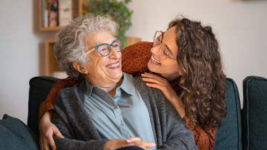 Older adult women with glasses and curly gray hair being embraced by a younger woman adult with curly brown hair.
