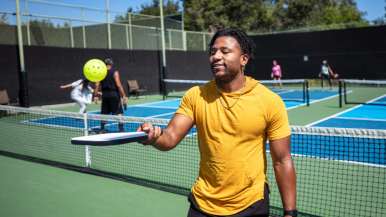 A young man bounces a pickleball on his paddle while a group plays a doubles match on the court in the background.