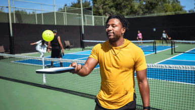A young man bounces a pickleball on his paddle while a group plays a doubles match on the court in the background.