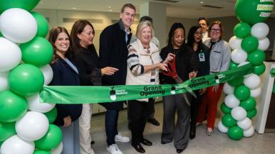 Group of people cutting a green ribbon at a grand opening event, surrounded by green and white balloon columns.