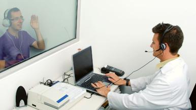 An audiologist conducts a hearing exam on a patient seated in an audiometric booth