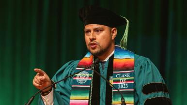 Jordan Cisneros, MD, speaks into a microphone and makes a pointing gesture with his right hand. He wears graduation regalia including a stole that says Clase de 2023