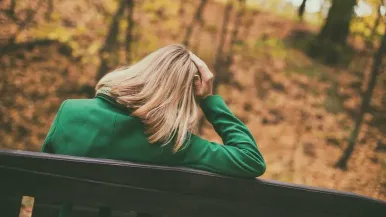 A woman in a green jacket sits on a park bench in autumn holding her head.