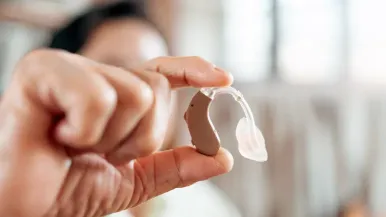 An older adult holds a behind-the-ear hearing aid to the viewer between thumb and forefinger.