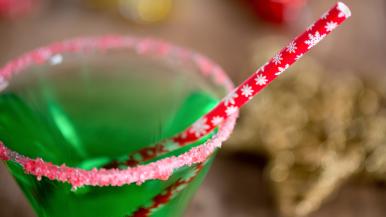 A martini glass rimmed with red and white sugar and filled with a green holiday drink. A red straw with a snowflake pattern sticks out of the glass.