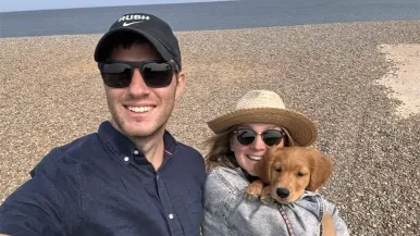 Ned Barker stands on a beach with his partner, Mandy, and their puppy.