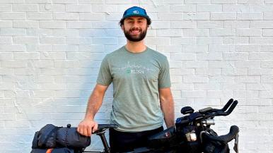 A smiling man wearing a cap and RUSH t-shirt standing beside a bike